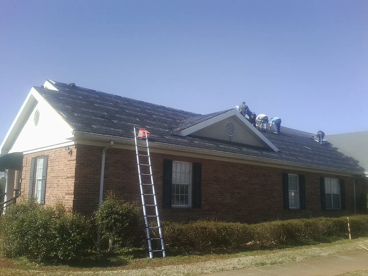 Crew tearing shingles off a brick ranch roof with a ladder set — Emergency Roof Repair work by our crew in Holly Hill