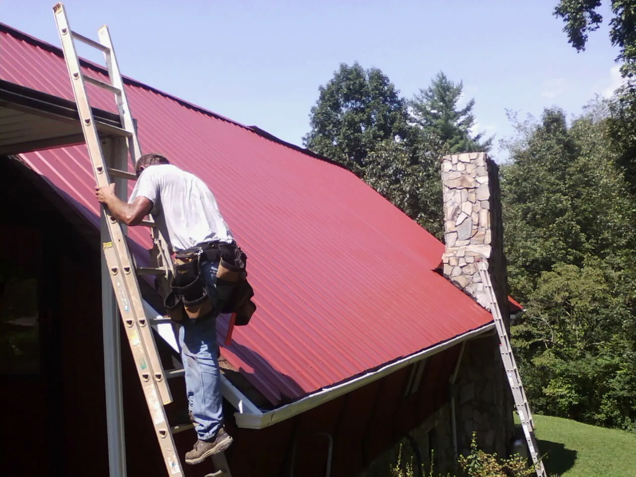Roofer on a ladder beside a red metal roof and stone chimney — Soffit & Fascia Repair by Roofing Brothers in Holly Hill